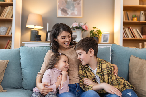 In their cozy living room, a mother sits on the sofa with her daughter and son beside her. They share everyday moments of laughter and conversation, basking in the simple joys of family togetherness.