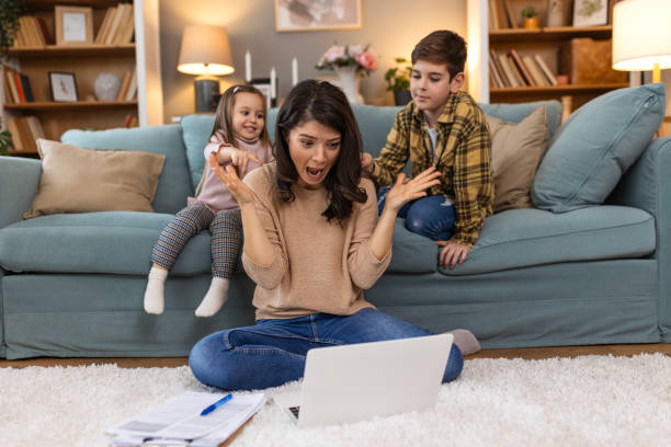 istock Tired young mother sitting on the floor and working with laptop and documents while little kids and jumping on the sofa and having fun and making noise 2062377094