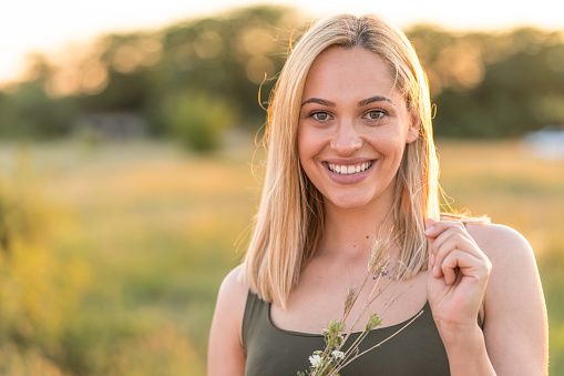 Portrait of beautiful blond woman holding a grass straw, looking at the camera in front of grass field during sunset