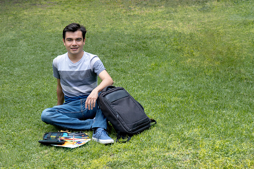 Young student man sitting on the grass with a backpack and school supplies