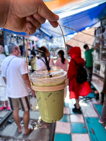 Maeklong Market, Bangkok, Thailand - February, 16 2024: Stock photo showing  close-up view of a lidded, plastic disposable cup with green drinking straw containing a green, matcha tea frappuccino drink held by unrecognisable person. This drink is made by blending ice, milk, vanilla syrup and green tea powder.