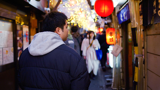 A medium close up shot from the back of young Asian man exploring Tokyo at night, walking down an alley in Omoide Yokocho in Shinjuku past izakaya restaurants.