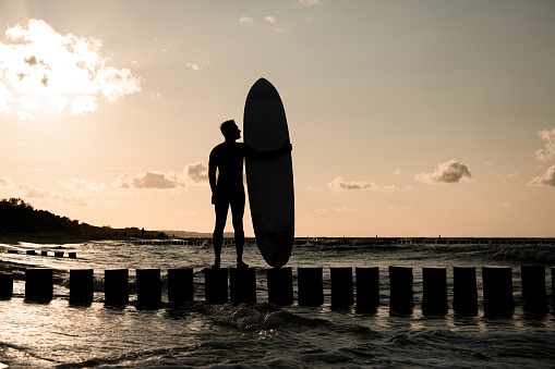 Young slim male surfer looking at his surfboard while standing on wooden stumps against a background of rough sea and crimson sky