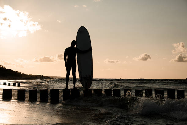 Young slim male surfer hugging his surfboard while standing on wooden stumps against crimson sky background