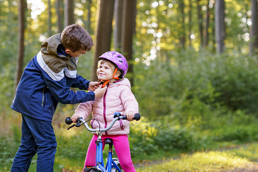 School kid boy, brother put on little preschool sister girl bike helmet on head. Brother teaching happy child cycling and having fun with learning bike. Active siblings family outdoors. Kids activity.