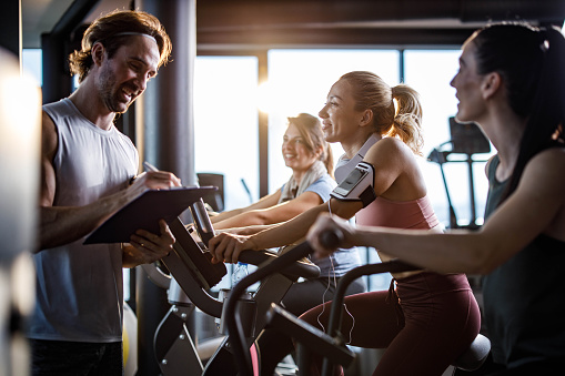 Happy woman exercising on stationary bike in a gym and looking at her coach writing plans for her future sports trainings.