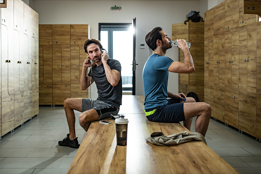 Two athletic men sitting on a bench in dressing room. One of them is listening music over cell phone while the other is drinking water.