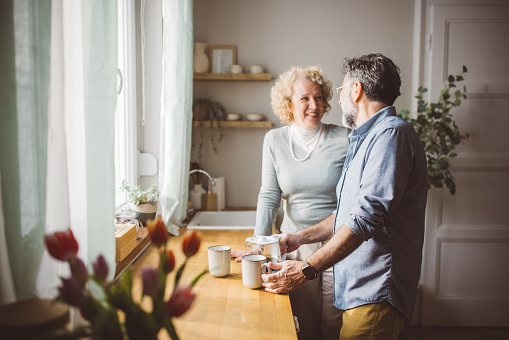 Mature couple standing in front of kitchen window and drinking tea or coffee. Enjoying in morning.