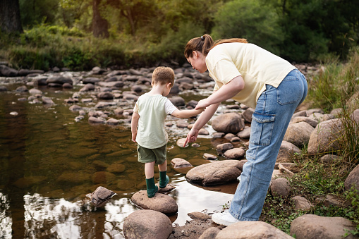 A mother helps her little son carefully cross natural stepping stones in a shallow mountain river.