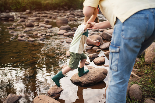 A mother helps her little son carefully cross natural stepping stones in a shallow mountain river.