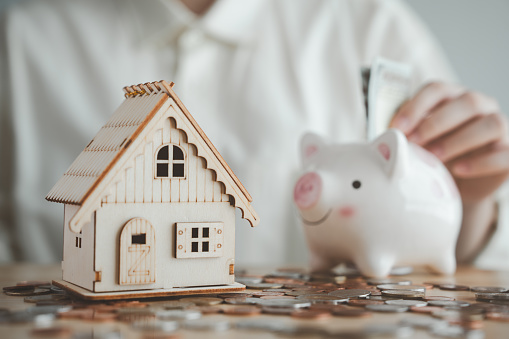 House model and hands are depositing money into a piggy bank. Plan to save money for a home budget, residential tax, home loan and mortgage rates. Concepts of business investment and real estate.