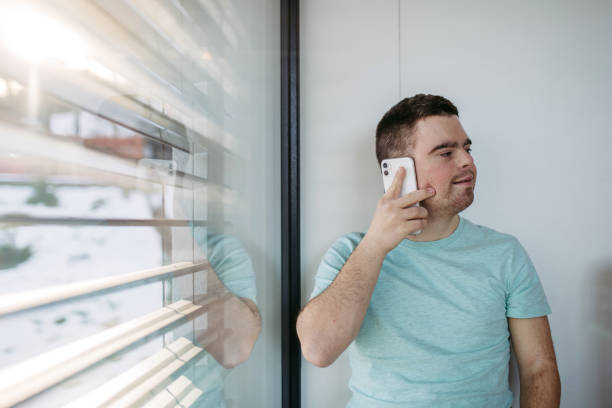 istock Young man with Down syndrome sitting by window, making phonecall with smartphone. 2062613055