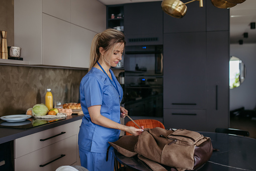 Female nurse, doctor getting ready for work, packing backpack, leaving house in scrubs with backpack. Work-life balance for healthcare worker.