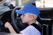 Little boy in a blue cap and with school bag sitting on the driver seat in a car. Little boy wants to start the car.