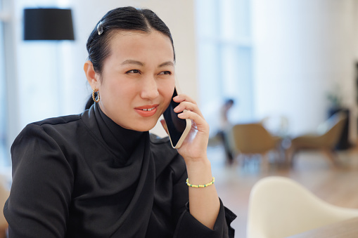 Beautiful young Asian woman working outside the office, sitting at hotel reception counter, using mobile phone and negotiating business, remote work