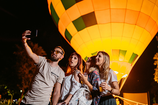 Group of young people taking selfies using smart phone in front of a hot air balloon at the music festival.