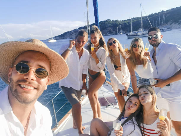Group of young people spending some great time together on sailing vacation Group of beautiful young people having a party on sailboat deck before sunset. They are dancing, drinking wine and cocktails, relaxing and enjoying their summer vacation on sailing yacht. One of them is using mobile phone to make a selfie with everybody. yacht selfie stock pictures, royalty-free photos & images