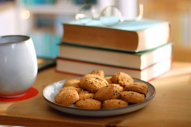 Cup of tea or coffee, pile of books, plate of cookies, reading glasses, e-reader and pen on the table. Colorful rainbow bookshelf in the background. Selective focus.