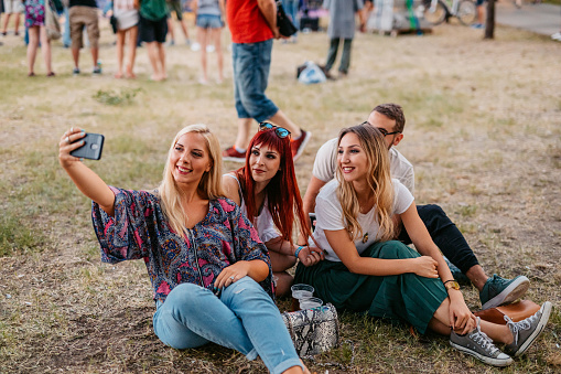 Group of young people sitting on the grass and taking selfies using smart phone at the music festival.