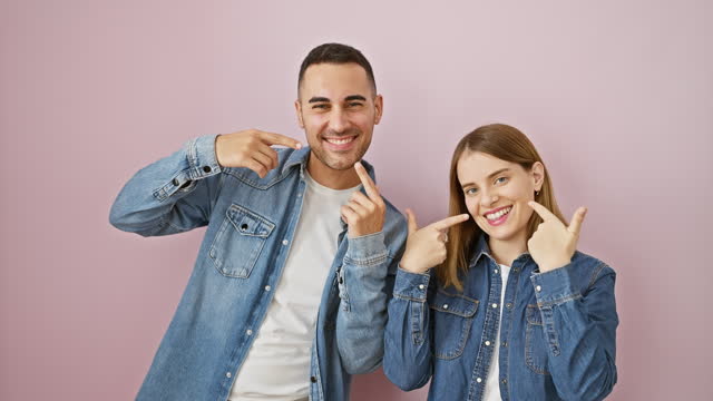 Cheerful beautiful couple, pointing at sparkling teeth with love, showcasing dental health over pink, isolated background, clad in denim shirts.