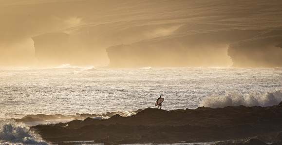 A surfer standing on large rocks by the water