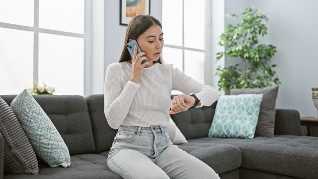 Hispanic woman checking time while talking on smartphone in a modern living room.