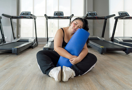 A plus size woman sit on the ground holding a blue exercise mat, resting during her workout.