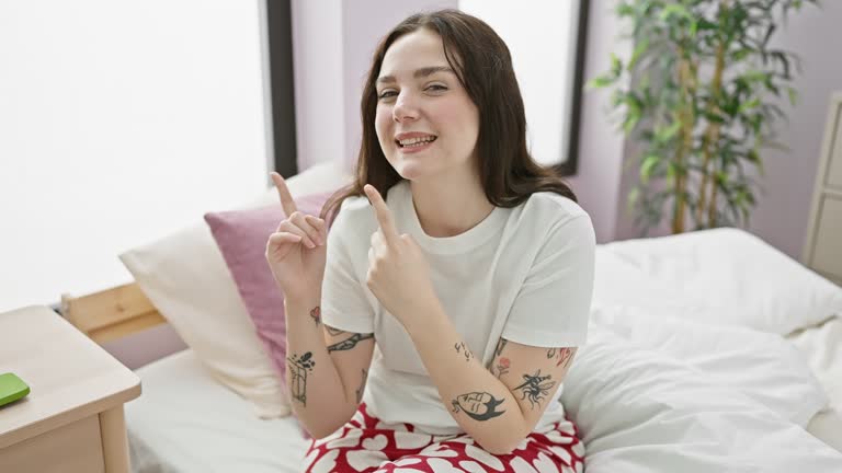 Cheerful young woman wearing pyjamas, sitting on bed, smiling and confidently pointing off to the side, inviting you into her cozy bedroom sanctuary.