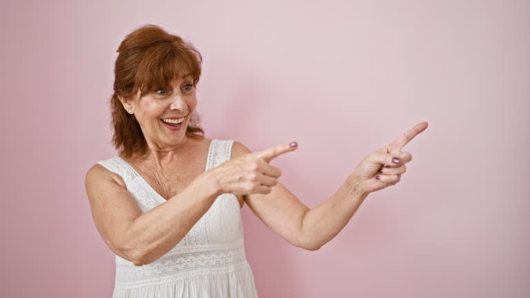 Cheerful middle-aged lady in dress, trustingly points and smiles at camera against pink wall