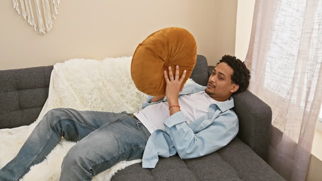 A relaxed man with curly hair lounging on a couch at home, holding a cushion, creating a cozy atmosphere.