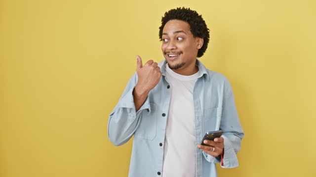 Joyful young african american man, confidently holding his smartphone, cheerfully points to the side with a thumbs up, standing against an isolated yellow background