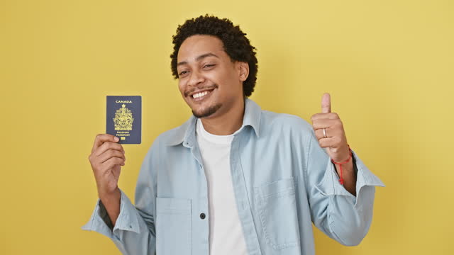 Joyful young african american man confidently showing off his canadian passport, giving the cool 'okay' sign; isolated on a cheerful yellow background