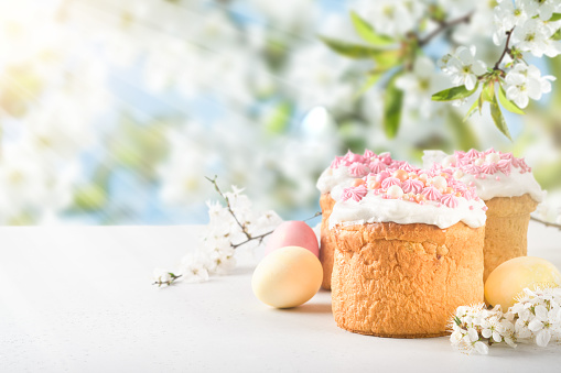 Traditional Easter cake with white swiss meringue and pink sweet decoration and colorful painted eggs on background blooming cherry or apple garden flooded with sunlight and rays with copy space.