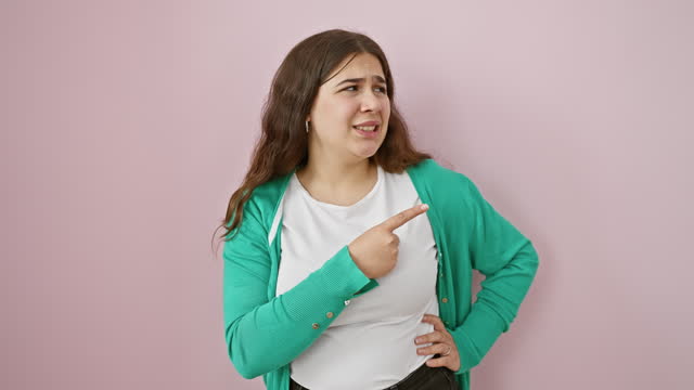Worried young hispanic woman with a beautiful nervous expression, standing over isolated pink background pointing aside with forefinger, surprise and concern painted on her face