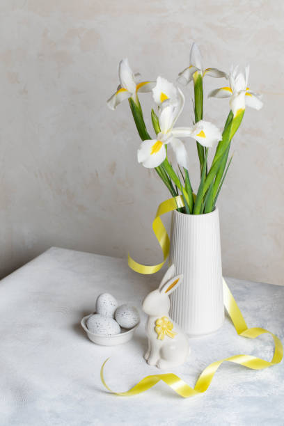 istock White Iris flowers bouquet in vase, decorative eggs, ceramic bunny on white table near white wall. 2062942056