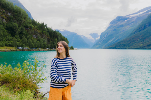 Handsome smiling female with long hair wearing stripe shirt and orange shorts admiring crystal blue lake Lovatnet with view of green mountain peaks and glacier in Jostedalbreen National park, Norway