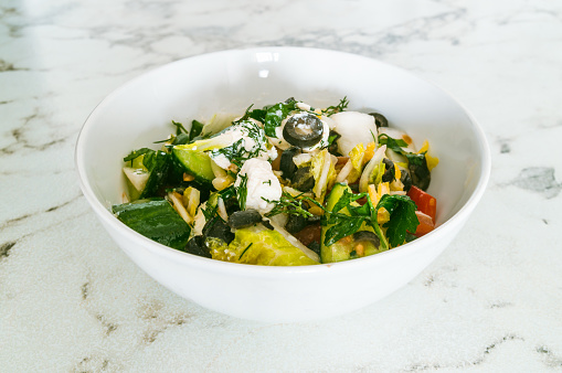 A white bowl filled with a salad of vegetables including cucumbers, tomatoes, and olives. The bowl is placed on a marble countertop