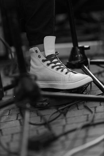 A black and white image capturing a musician foot adorned in a white sneaker, surrounded by intertwined cables on a stage floor. The contrast between the worn sneaker and the stage setting creates a captivating visual.