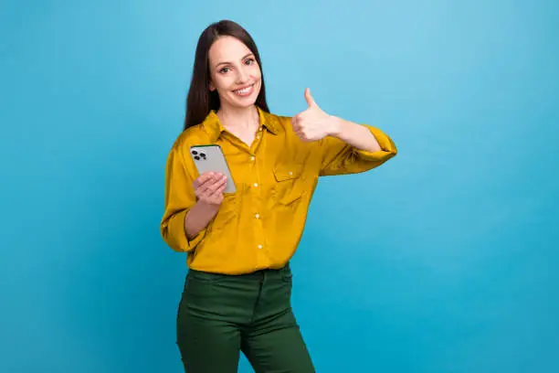 Photo of shiny confident girl dressed yellow shirt showing thumb up chatting modern gadget empty space isolated blue color background.