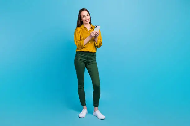 Full length photo of sweet shiny woman dressed yellow shirt communicating modern gadget empty space isolated blue color background.