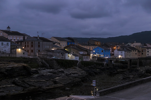 coastal town at dusk, with illuminated buildings nestled against a backdrop of darkening skies and rugged terrain