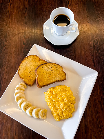 a white square plate for breakast with eggs, bananas and toast bread on a wooden table background