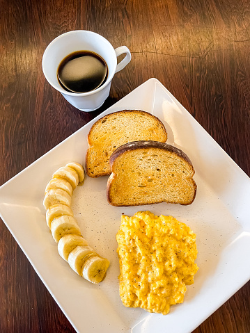 a white square plate for breakast with eggs, bananas and toast bread on a wooden table background