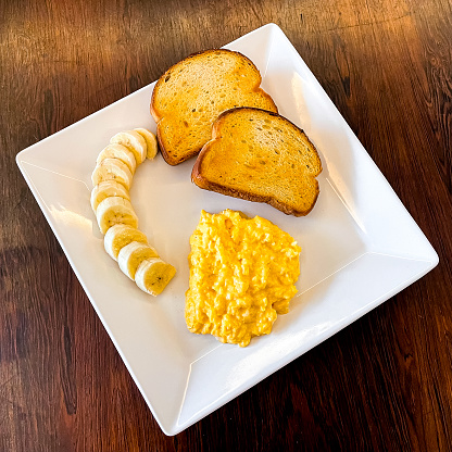 a white square plate for breakast with eggs, bananas and toast bread on a wooden table background