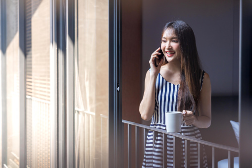 casual relax asian woman in casual cloth dress drinking cup hot coffee while talking on smartphone clam peaceful lifestyle peaceful stay in modern home architecture home design view from outside window bedroom