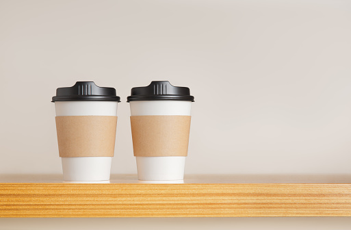 Two paper coffee cups stand on the bar on beige background. Close-up and copy space.