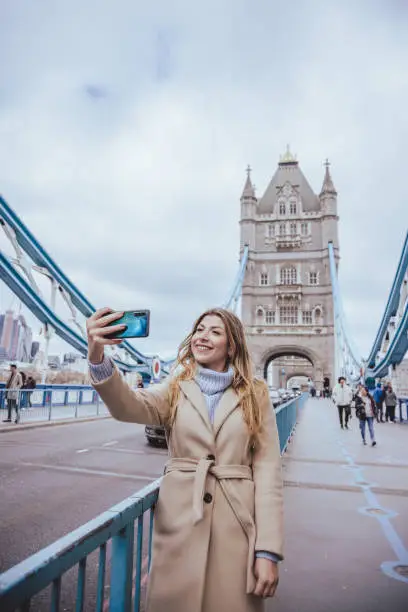 Candid portrait of young blond woman in her 30s sightseeing London's iconic landmarks, smiling and taking selfie with a smart mobile phone device(not looking into the camera) at Tower Bridge in city of London, United Kingdom. Image created during daytime, outdoors an a cloudy day during cold season with warm casual clothes in light cream and blue colours. Vertical shot with plenty of copy space, created with wide angle lens used to capture the surroundings including accidental unrecognisable tourists and traffic passing over the iconic structure -  creative stock photo