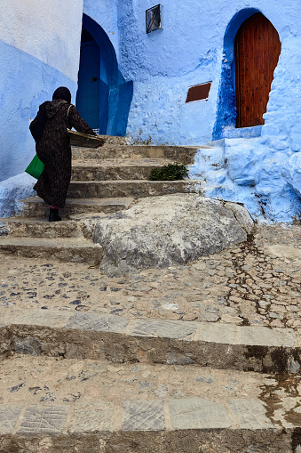 Woman in black djellaba walking upstairs view from behind in blue city, Chefchaouen, Morocco, North Africa.