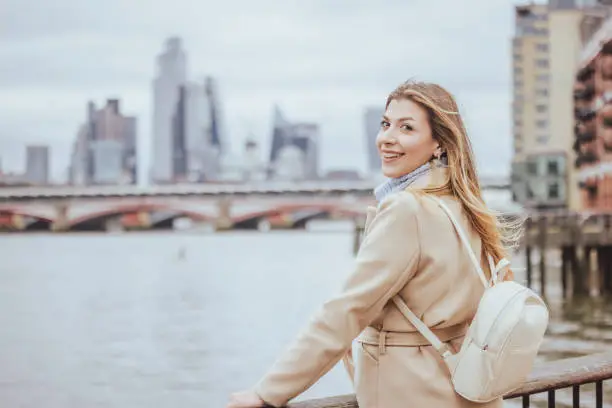 Waist up portrait of a young cheerful blond woman in her 30s posing and smiling in toothy smile, while looking directly in camera, the background is city of London's skyline, England, UK. Selective focus with priority on the model and plenty of copy space at the background and sky, which is defocused down town modern corporate business buildings and financial district near Liverpool Street at the heart of the capital. Photo created during cold season outdoors and the model is with warm casual clothes in light cream and blue colours on a cloudy day  - creative stock photo