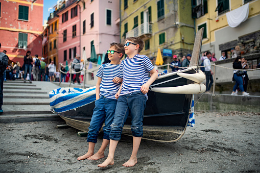 Family sightseeing beautiful Italian town of Vernazza.  One of the five towns in Cinque Terre National Park - a UNESCO World Heritage Site. Two little boys are leaning against a fishing boat in the harbor, enjoying a break from sightseeing on the small beach.\nShot with Nikon D850